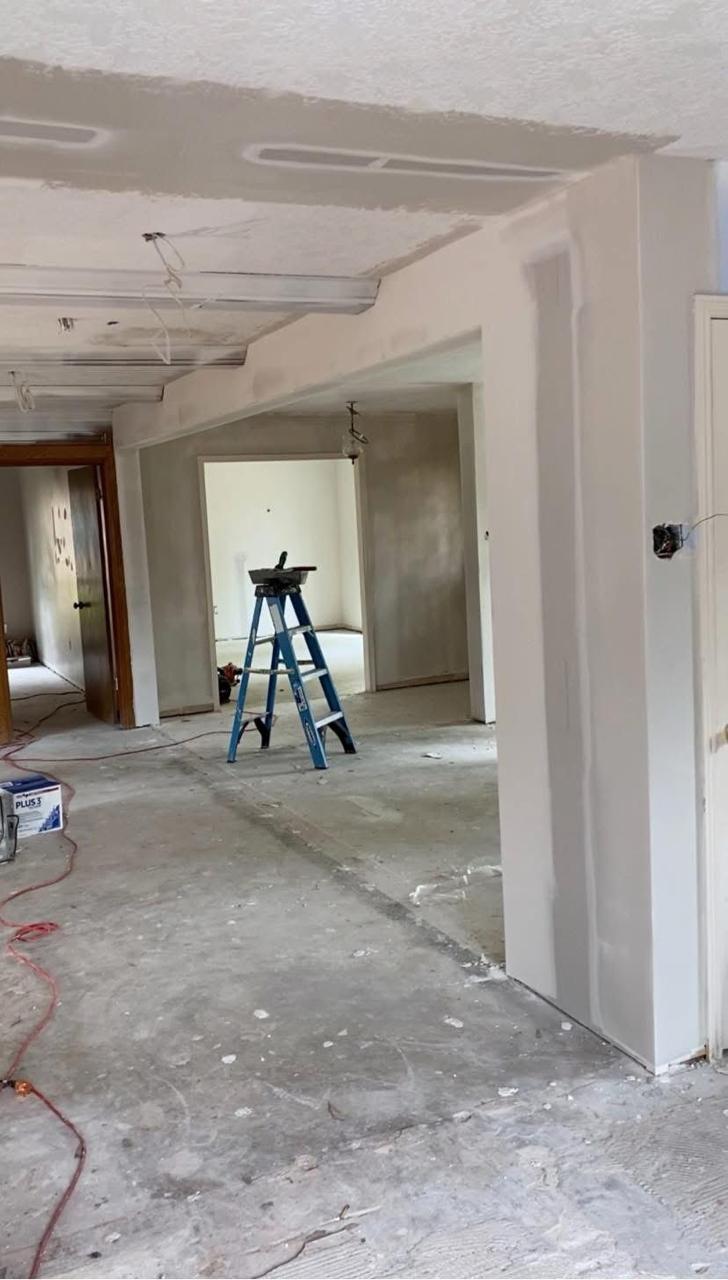 Open plan interior before — drywall finishing in progress, exposed flooring, ladder, and ceiling prep
